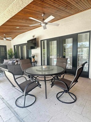 An outdoor patio with a round glass table, four chairs, a ceiling fan, and a TV mounted on the wall. - Villa Van Vista Luxury Gulf Access Retreat (Cape Coral)