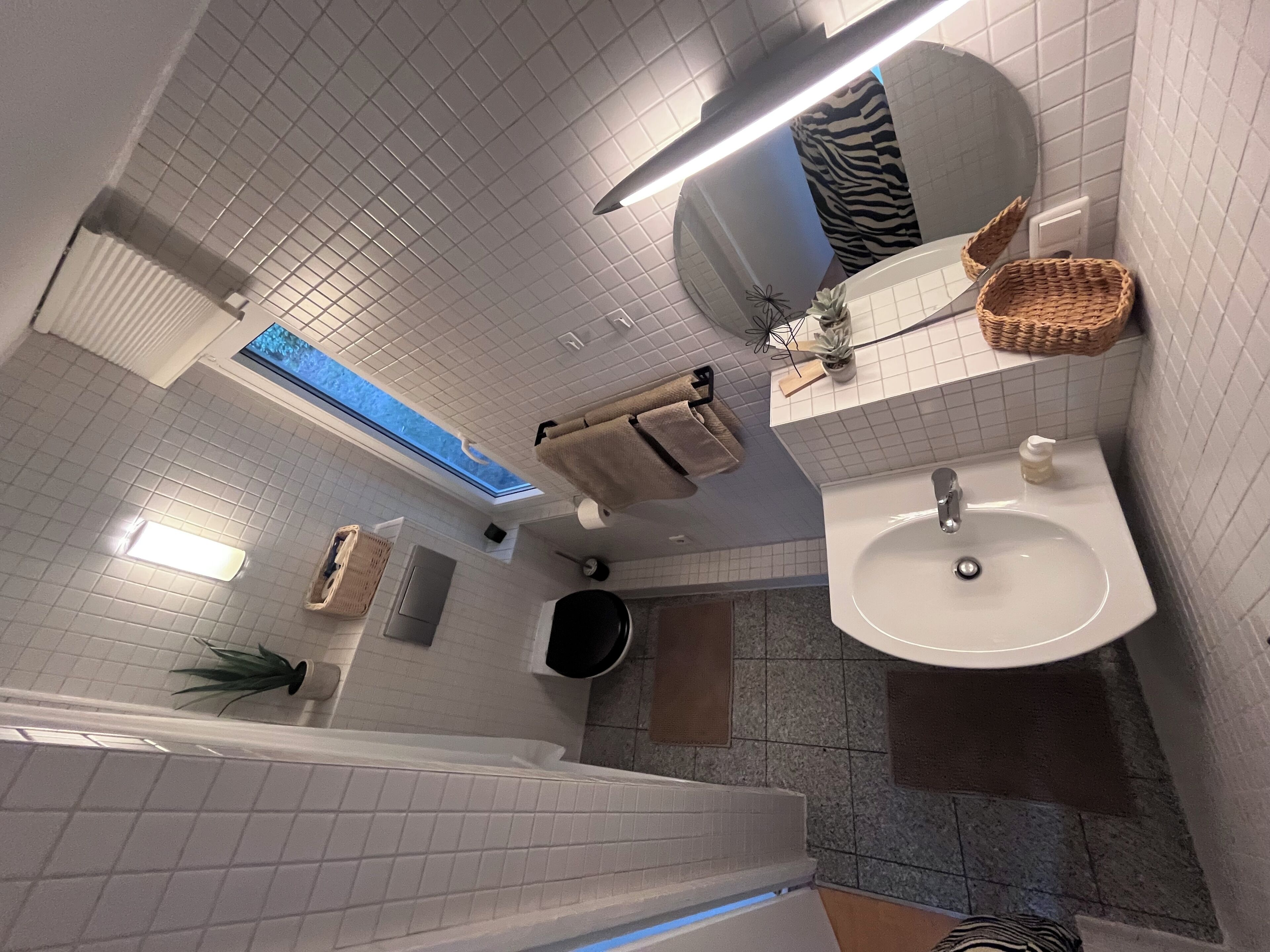 Ein modernes Badezimmer mit Oberlicht, großem Spiegel, Waschbecken und Duschbereich.