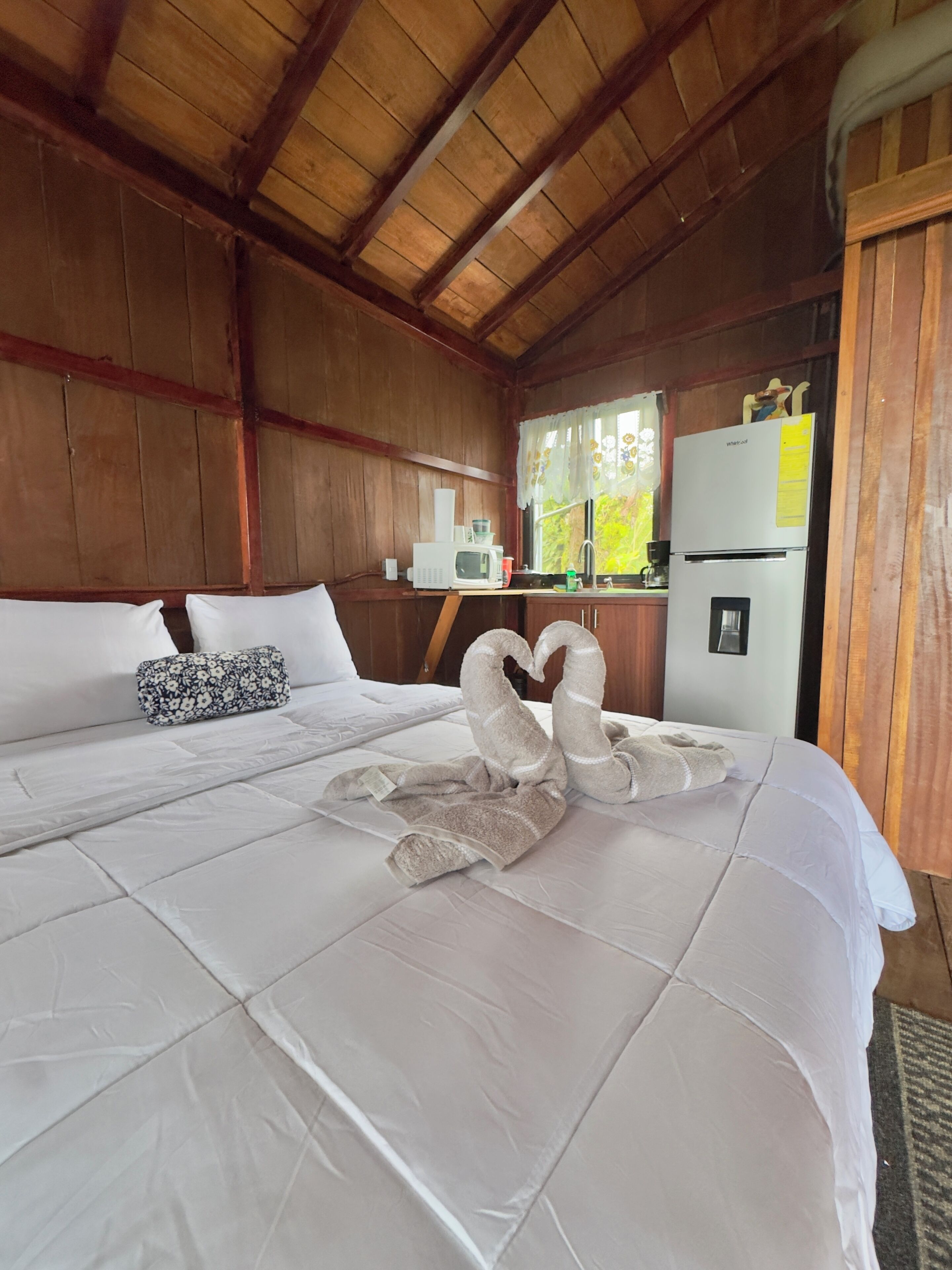 Habitación de hotel con techo de madera, cama con ropa de cama blanca, refrigerador blanco y ventana con vistas a zonas verdes.