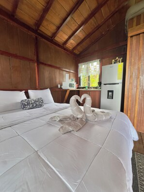 A hotel room with a wooden ceiling, a bed with white linens, a white refrigerator, and a window with a view of greenery. - Mountain Cabin | Sloths | Panoramic View (San Carlos, La Tigra)