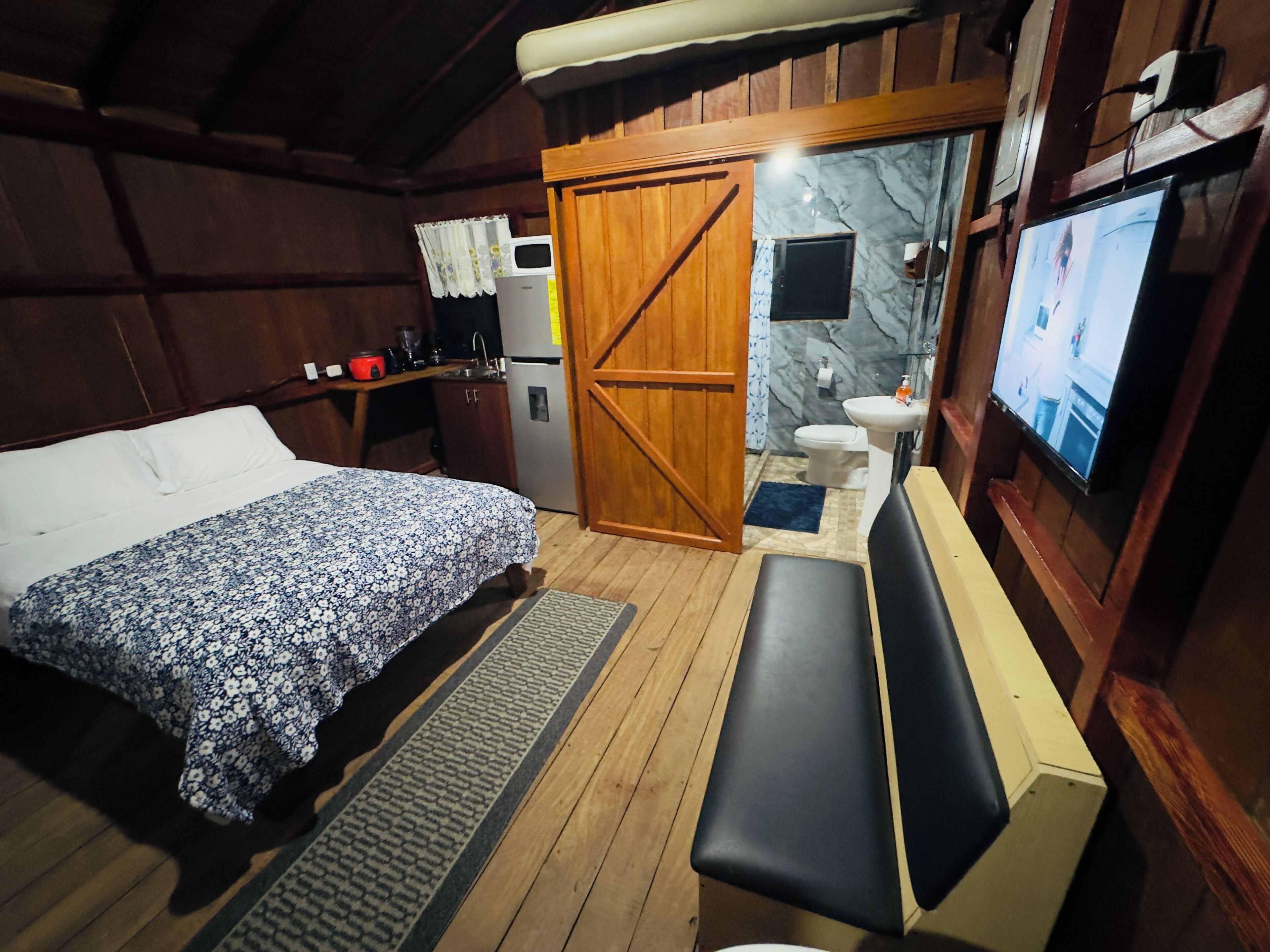 Una habitación con cama, un banco, televisión, lavamanos y un inodoro.