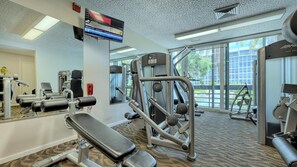 Ein Fitnessstudio mit verschiedenen Trainingsgeräten, einem großen Spiegel und einem Fernseher, der Finanznachrichten zeigt.
