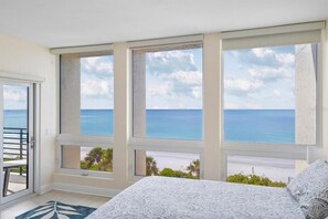 A bedroom with a large window overlooking a beach, a bed with patterned bedding, and a door leading to a balcony. - Stunning Beachfront 2 BR Condo (Longboat Key)