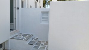 Un couloir extérieur étroit, avec un mur blanc, une porte et une petite fenêtre.