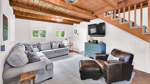 Uma sala de estar ampla com um sofá cinzento, uma poltrona de couro marrom, um aparador azul e uma escada de madeira.
