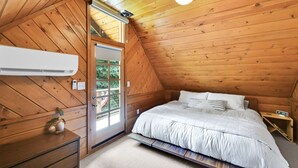 Quarto com teto inclinado, uma cama com roupa de cama branca, uma mesinha de cabeceira e um ar condicionado fixado na parede.