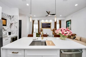 A modern kitchen with a central island, pendant lights, a ceiling fan, and a dining area with a sofa and decorative flowers. - Quiet Elegance• 2 Homes •NRG Stadium • Med Center • Sleeps 20+ Large Group Stays (Houston)