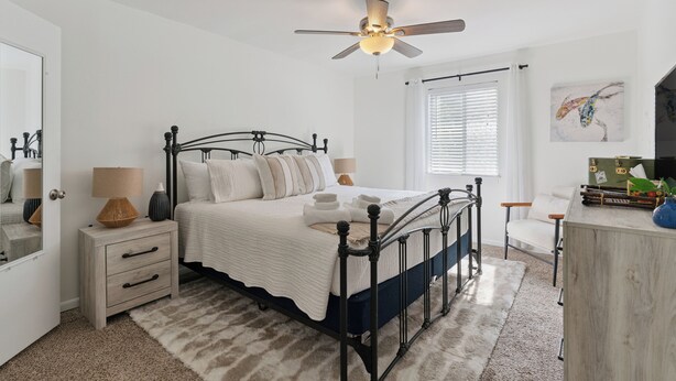 A bedroom with a bed, bedside tables, a nightstand, a ceiling fan, a window with blinds, and a painting on the wall. - The Elliott, Ocean Walk Getaway, Pools & Walkable Location (St. Simons Island)