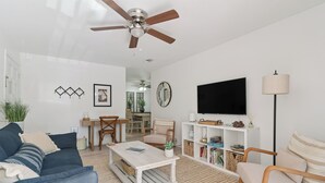 A modern living room with a ceiling fan, a flat-screen TV, a bookshelf, and a dining area with a table and chairs. - The Elliott, Ocean Walk Getaway, Pools & Walkable Location (St. Simons Island)