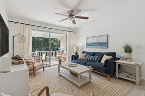 A spacious living room with a large window, a ceiling fan, a sofa, a coffee table, and a TV on a stand. - The Elliott, Ocean Walk Getaway, Pools & Walkable Location (St. Simons Island)
