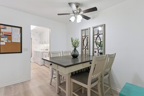A dining area with a table, chairs, a ceiling fan, and a bulletin board with papers. - The Elliott, Ocean Walk Getaway, Pools & Walkable Location (St. Simons Island)