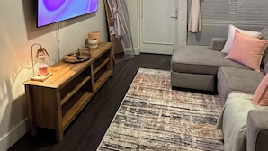 Una sala de estar con televisor de pantalla plana, ventilador de techo, banco de madera, sofá gris y alfombra estampada.
