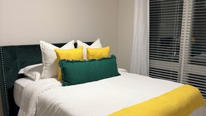 Cama con ropa blanca, almohadas verdes y amarillas, y una manta amarilla.