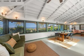 A spacious room with a high ceiling, large windows with blinds, a dining area with a table and chairs, and a green upholstered sofa. - 2BR West Maui condo with pool, patio, AC, and full kitchen -ICC 4 (Lahaina)
