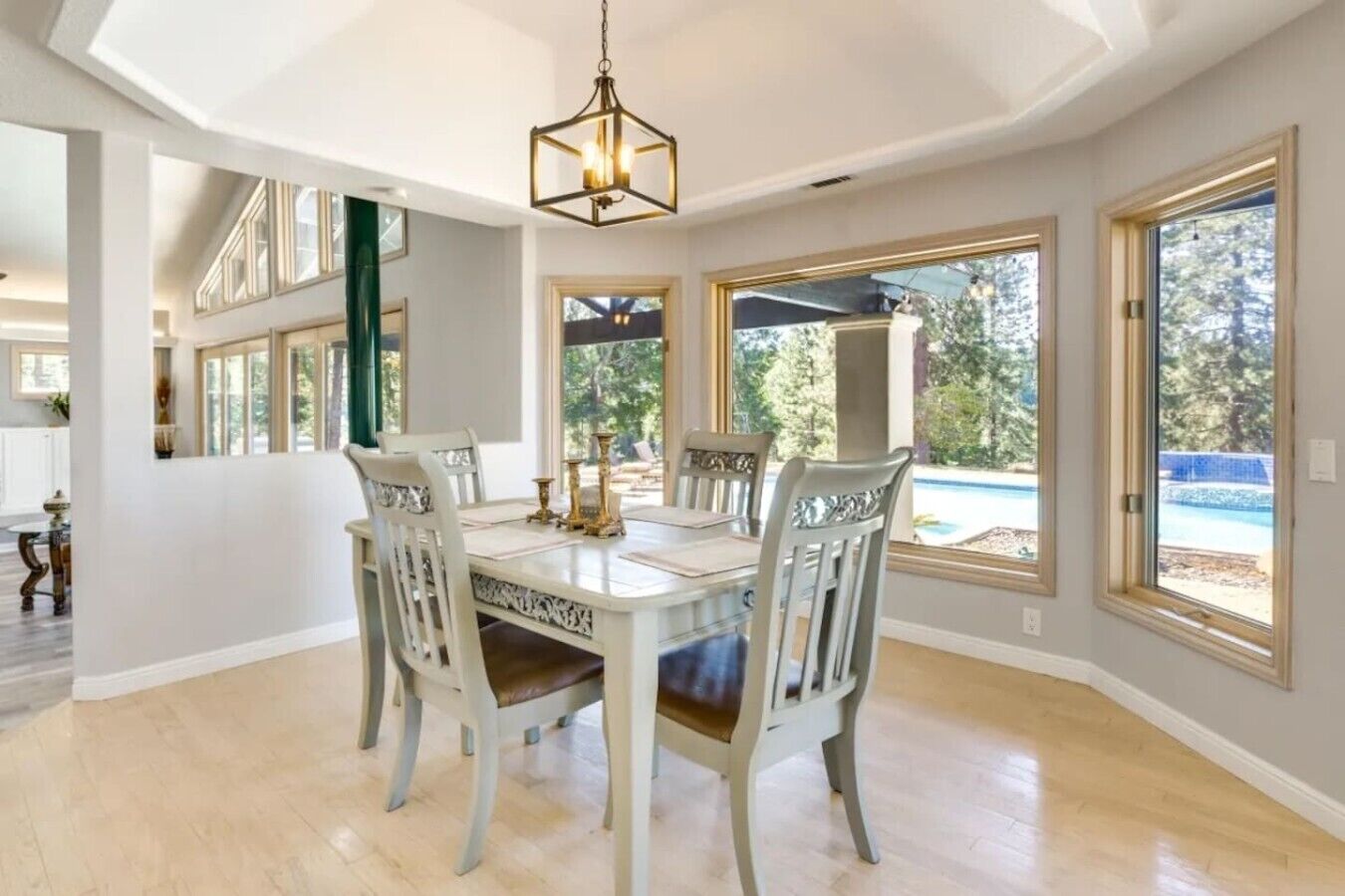 Un comedor con una mesa grande y sillas, ventanas amplias con vista a una piscina, y una lámpara colgante.