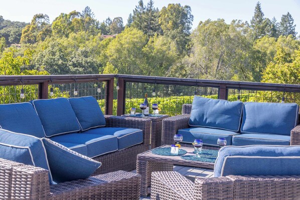 A covered outdoor seating area with wicker furniture and blue cushions, overlooking a forested landscape.
