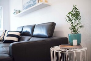 A modern living room with a dark leather sofa, a white round side table with a plant and a candle, and a white wall with a shelf. - BONDI HAVEN- CHIC 2 BEDROOM APARTMENT (Bondi)