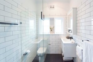 A modern bathroom with white subway tiles, a glass shower enclosure, a white sink, and a window with a plant. - BONDI HAVEN- CHIC 2 BEDROOM APARTMENT (Bondi)