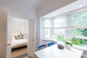A room with a bed, a dining table with chairs, and a window with blinds. - BONDI HAVEN- CHIC 2 BEDROOM APARTMENT (Bondi)