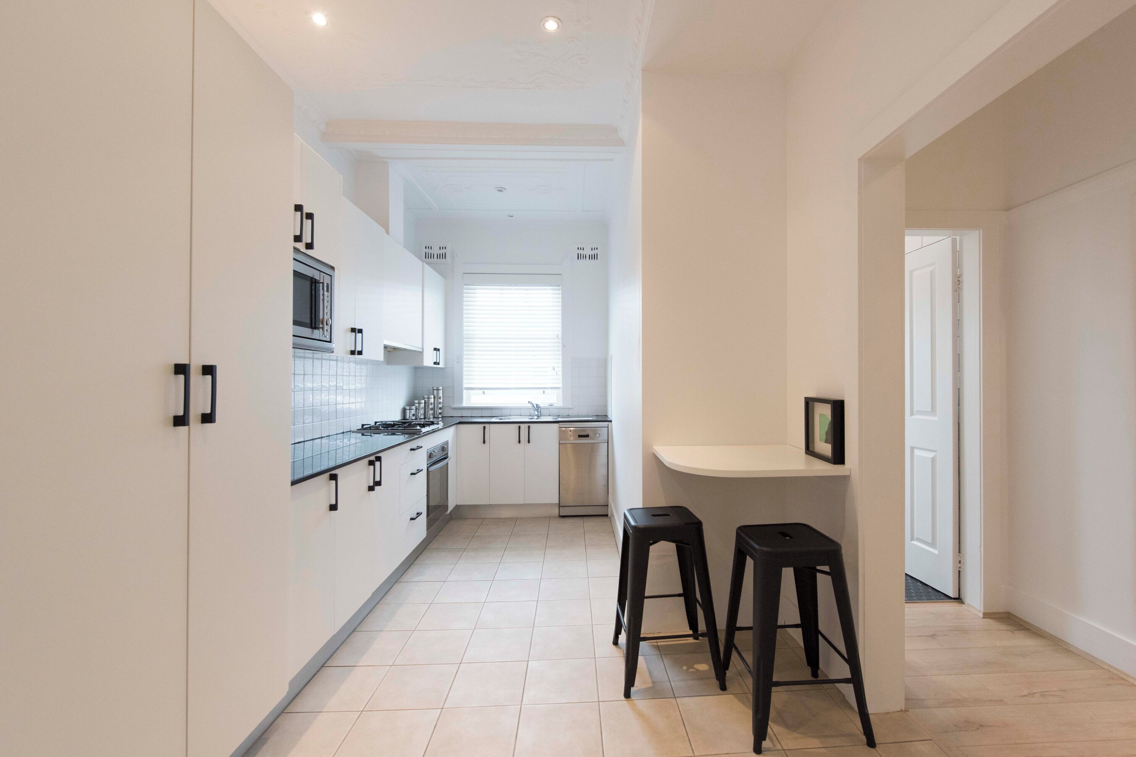 A modern kitchen with white cabinets, a microwave, stove, and a dishwasher. There are two black stools and a small white counter.