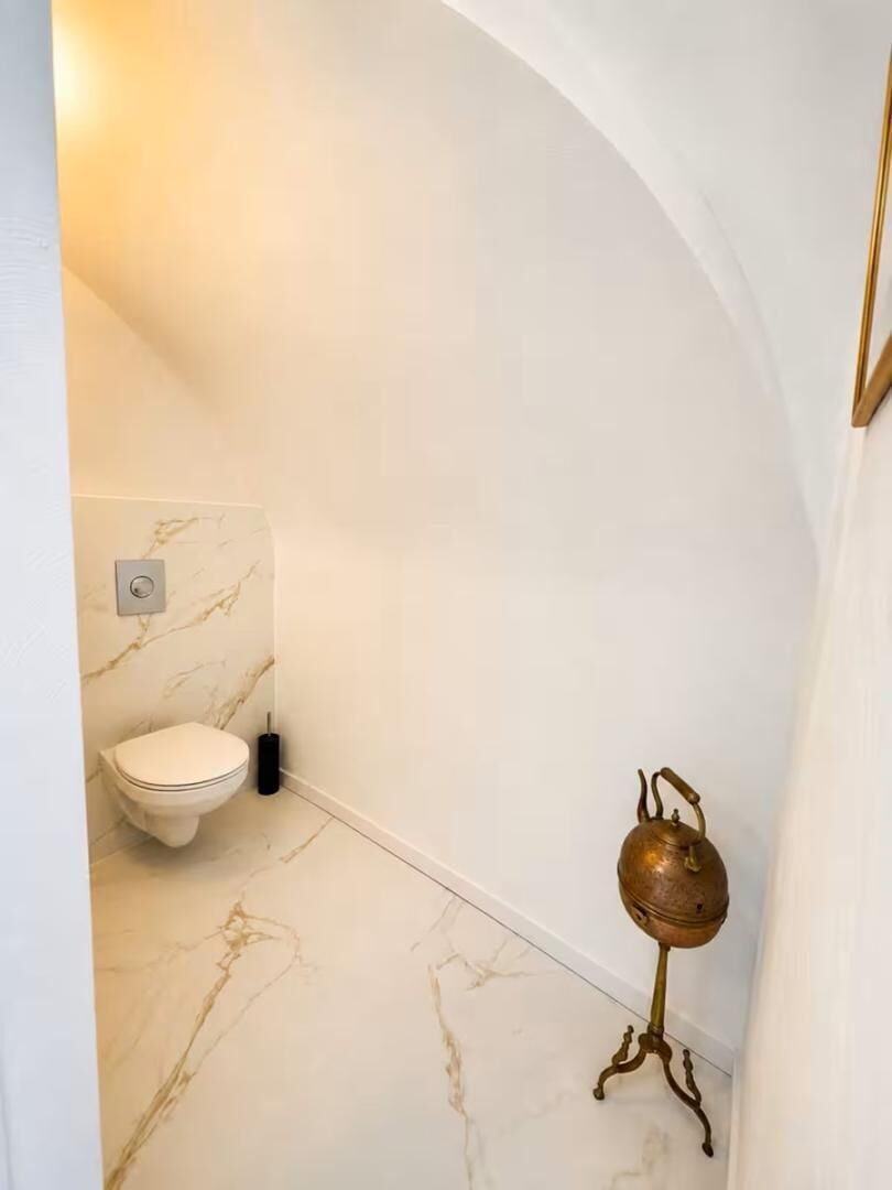 A bathroom with a marble floor and walls, a white toilet, and a brass kettle on a stand.