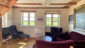 A room with wooden beams, a television, two chairs, a table, and two windows with views of greenery.