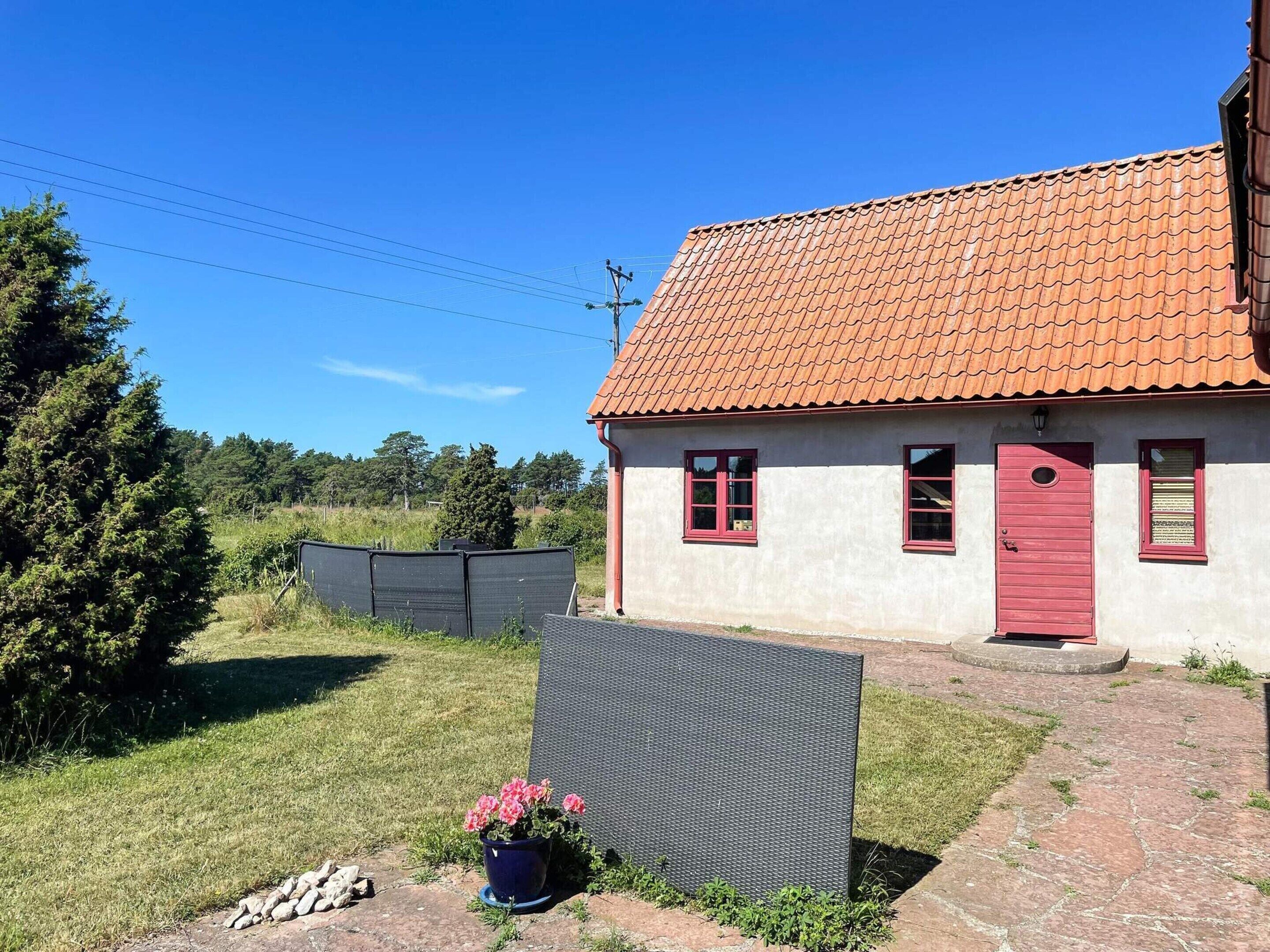 A single-story house with a red-tiled roof and a red door, surrounded by a grassy area and a metal fence.
