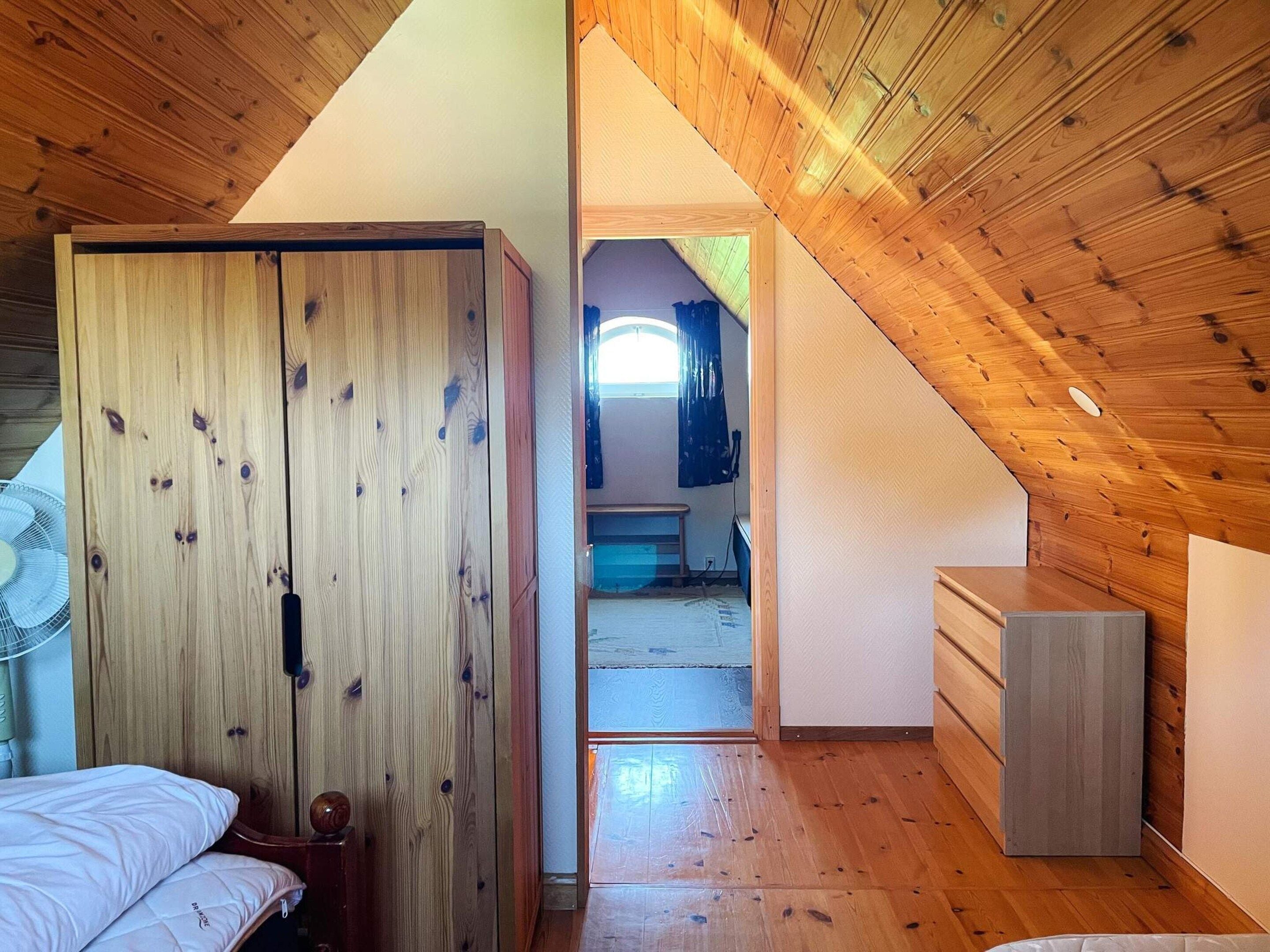 A room with a sloped ceiling, wooden walls, a bed, a dresser, and a fan.