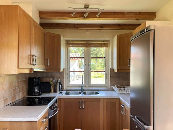 A kitchen with wooden cabinets, a stainless steel refrigerator, a built-in oven, and a window with blinds.