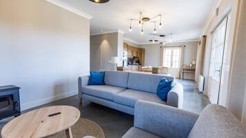 Een moderne woonkamer met een grijze bank, een ronde houten tafel en een keuken op de achtergrond.