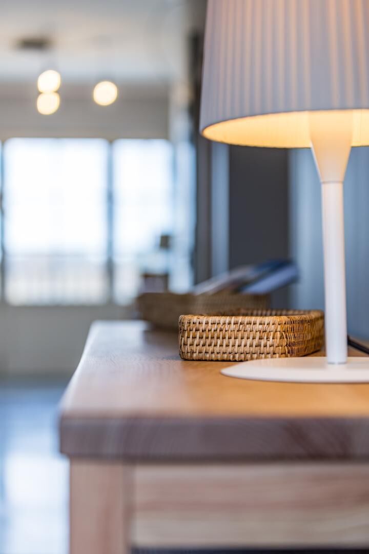 A modern interior with a lamp on a wooden table, a woven basket, and a blurred background suggesting a spacious room.