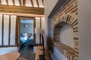 Ein Zimmer mit freiliegenden Holzbalken, einer Ziegelwand mit Bogen, einem Bett mit blauer Bettwäsche, einem Schreibtisch mit Lampe und Blick in einen weiteren Raum.