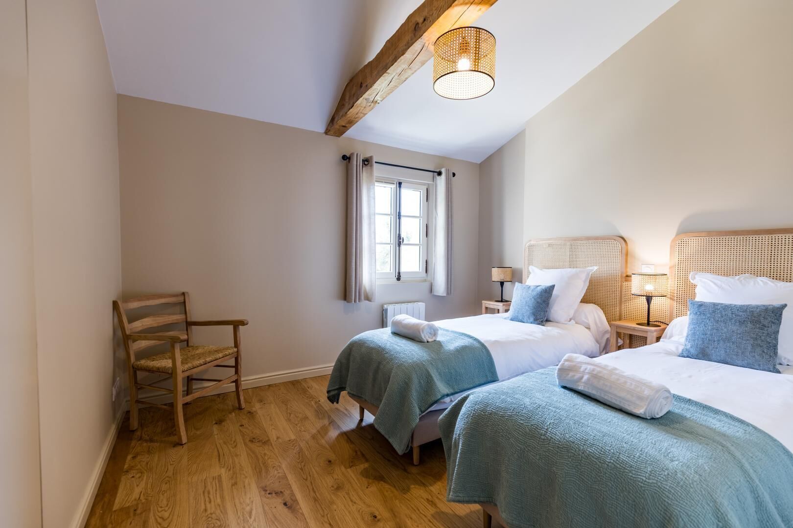 A bedroom with two beds, a wooden chair, a window with curtains, a wooden ceiling beam, and a hanging light fixture.
