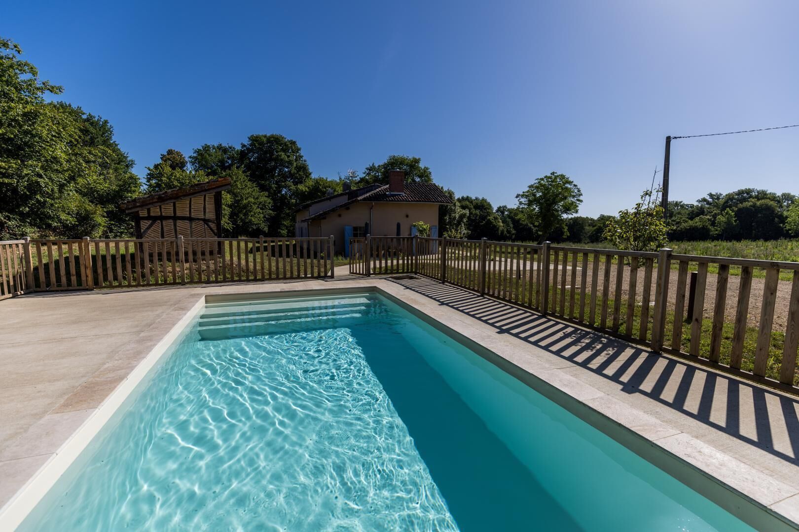 Une piscine rectangulaire à l’eau claire, entourée d’une clôture en bois, avec un bâtiment visible au loin.