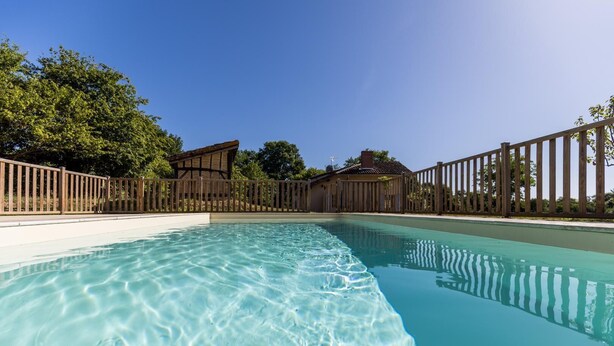Une piscine aux eaux cristallines, entourée d’une clôture en bois, avec un espace salon couvert et des arbres en arrière-plan.