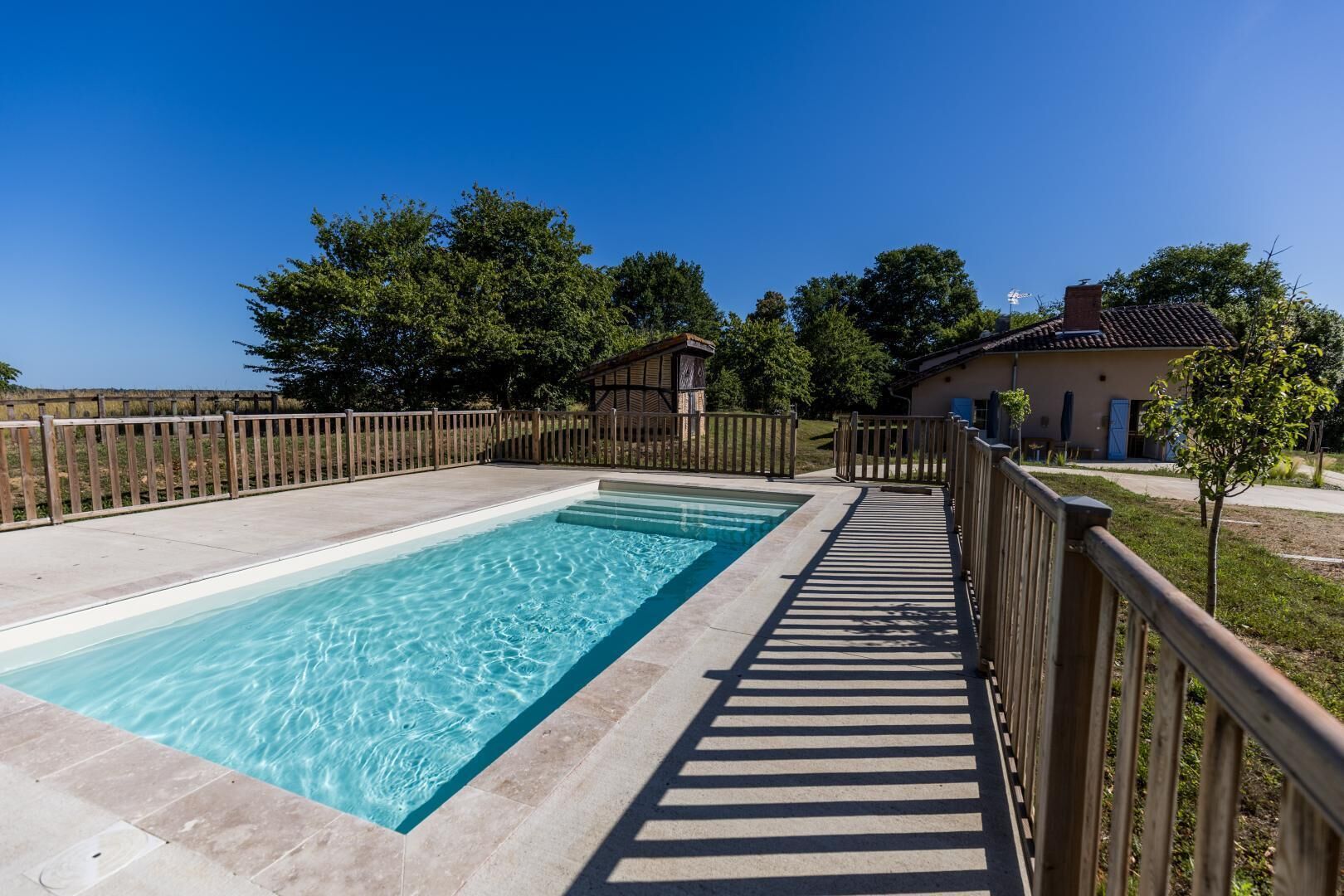 Une piscine rectangulaire à l’eau claire, entourée d’une clôture en bois et bordée d’un chemin pavé.