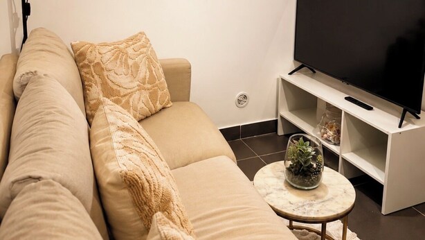 A modern living room with a beige sofa, a flat-screen TV, a small round coffee table, and decorative items on shelves.