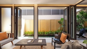 Una sala de estar moderna con un sofá, una mesa de centro y una silla. Hay un ventanal corredero que da a un balcón con vistas a una piscina y vegetación.