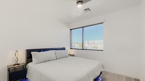 A bedroom with a bed, a nightstand, a lamp, and a window with a city view. - The Neon Circuit Loft (Phoenix)