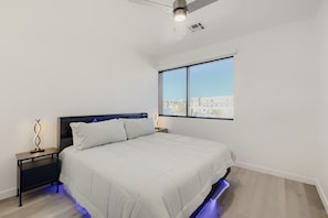 A bedroom with a bed, a nightstand, a lamp, and a window with a city view. - The Neon Circuit Loft (Phoenix)