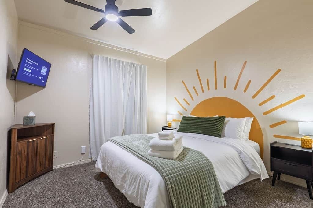 A hotel room with a bed, a ceiling fan, a television, a nightstand, and a wall with a sun design.