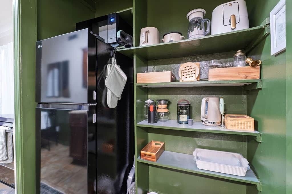 A kitchen with a black refrigerator, shelves with kitchenware, and a wooden cutting board.