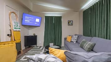 Un saló amb un sofà gris, coixins grocs i grisos, una televisió penjada a la pared, una cortina verda i una taula petita amb un llibre.