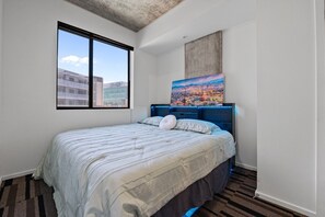 A hotel room with a large bed, a cityscape painting, and a view of buildings through a window. - Phoenix Scholars Loft (Phoenix)