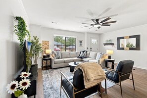 Ein modernes Wohnzimmer mit einer Couch, einem Couchtisch und Sesseln, einem Deckenventilator und einem großen Fenster mit Jalousien.