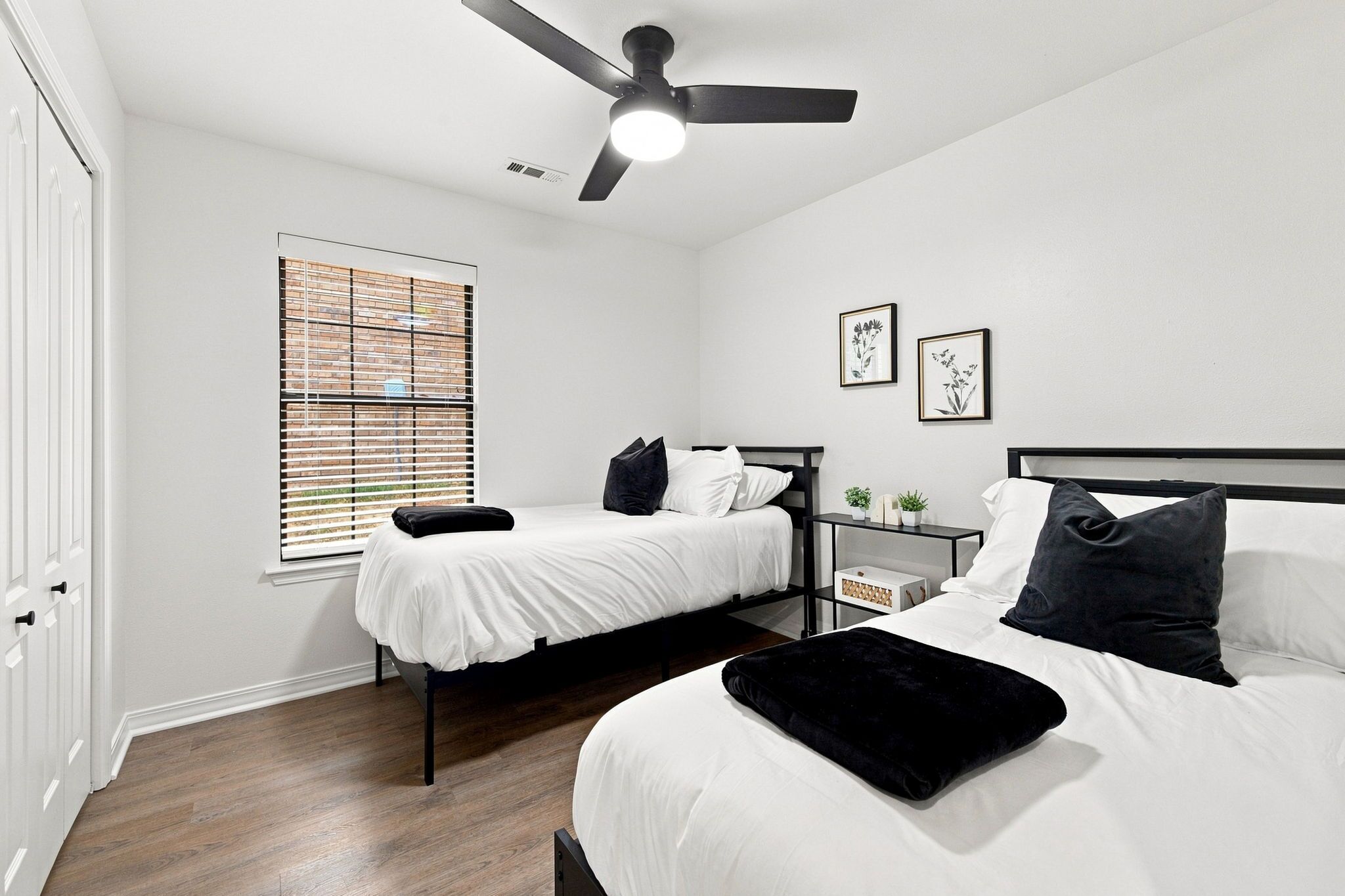 A bedroom with two beds, a ceiling fan, a window with blinds, and a bedside table with a plant.