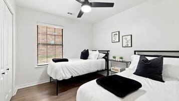A bedroom with two beds, a ceiling fan, a window with blinds, and a bedside table with a plant.