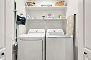 A laundry room with two white front-loading washing machines and a dryer, a broom, and a vacuum cleaner on the wall. - Switchback Suites - Side A (Bentonville)