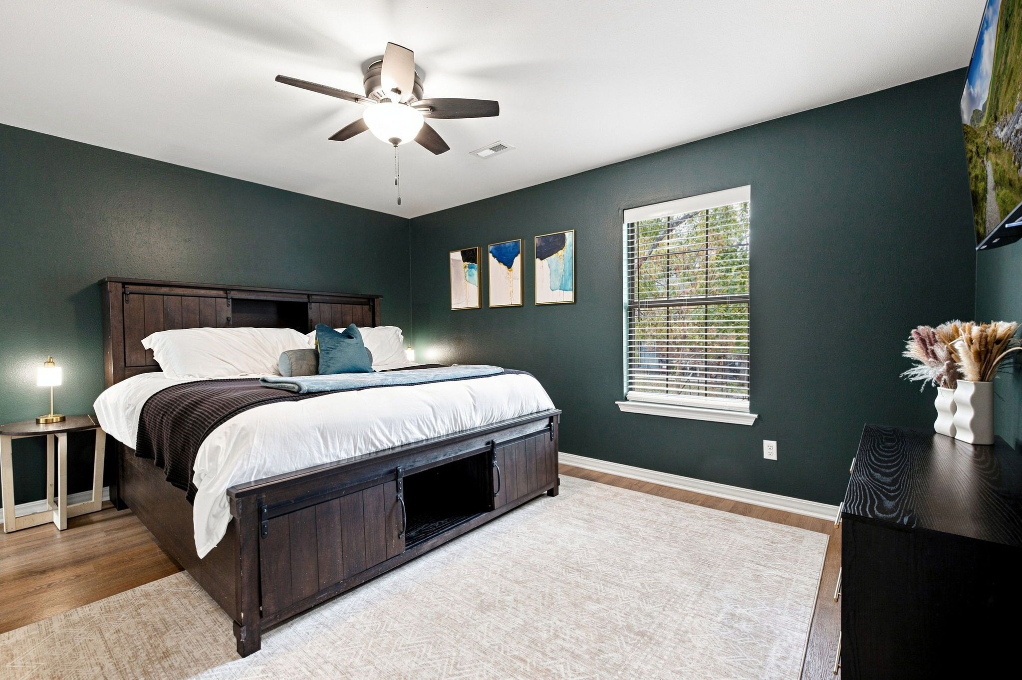 A bedroom with a bed, a ceiling fan, a window with blinds, and artwork on the wall.
