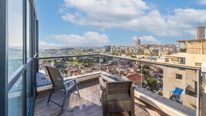 A balcony with a view of a cityscape, a wicker chair, and a wooden deck. - Premium and Luxe 2BR by the Shuk – Free Parking (Jerusalem)